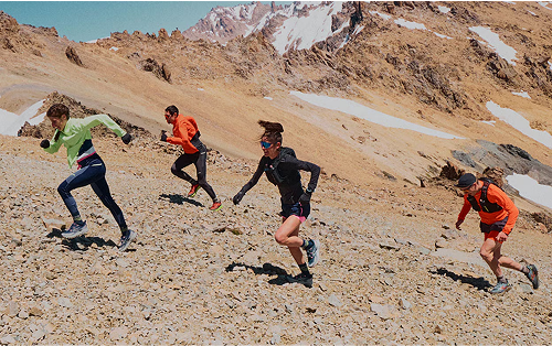 Quatro pessoas correndo em alta velocidade por um terreno rochoso nas montanhas, vestindo roupas esportivas coloridas. O cenário ao fundo mostra picos cobertos de neve sob um céu azul claro.
